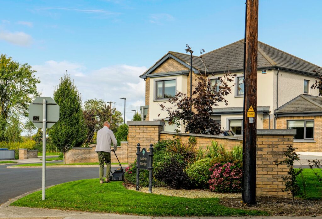 A man mowing the lawn outside a modern suburban home with a lush garden and clear blue sky.