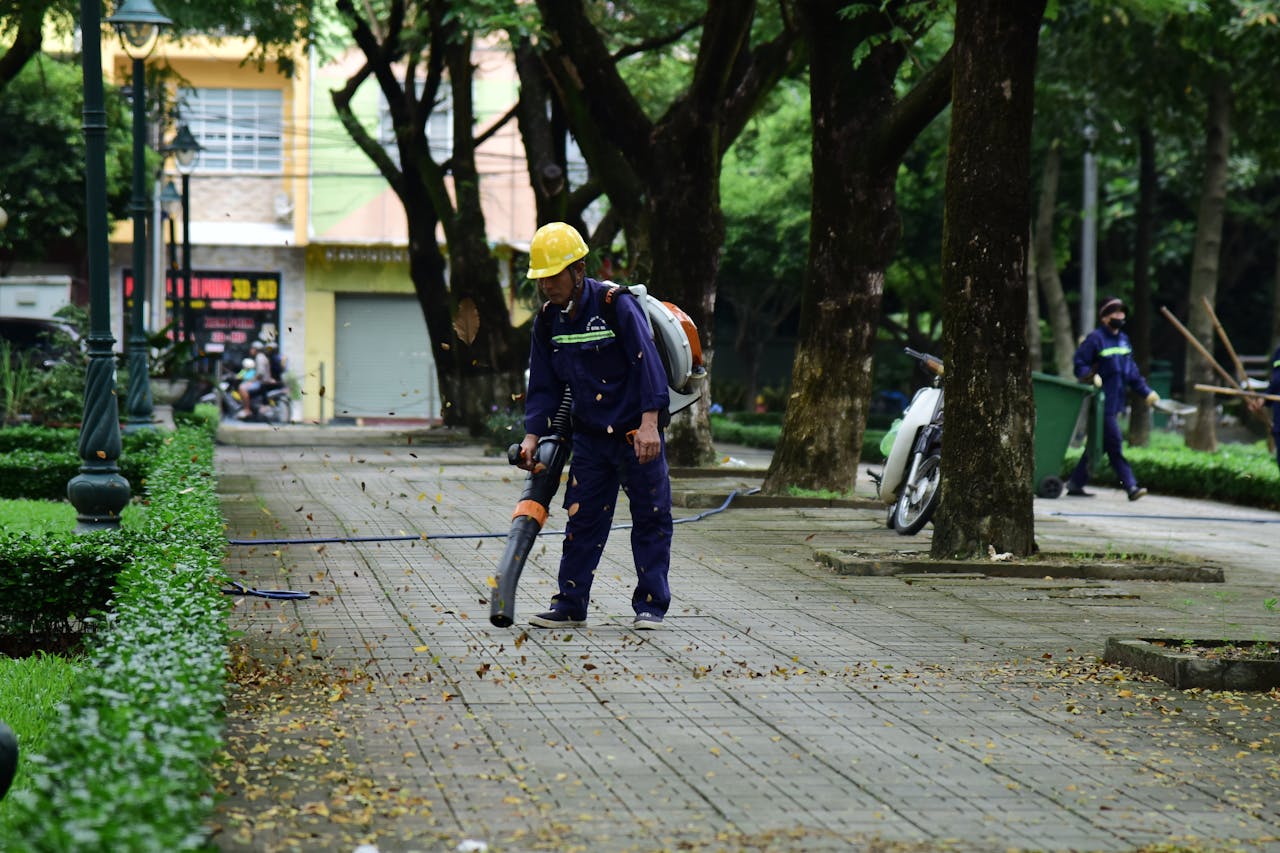 A city worker wearing protective gear uses a leaf blower to clean a public park pathway.